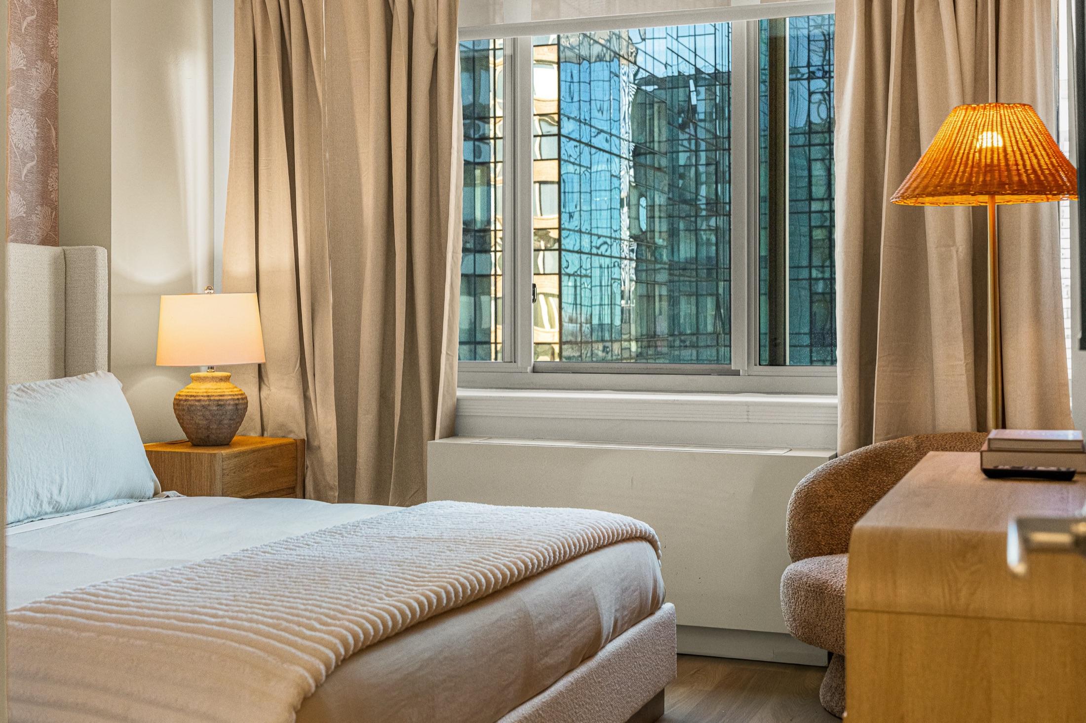 Short-term rental bedroom with floor-to-ceiling city views and warm lamp light, Manhattan
